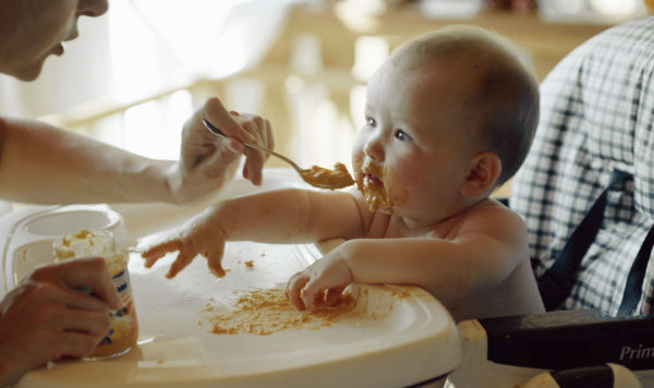 Мать кормит с ложки ребенка, сидящего за столиком