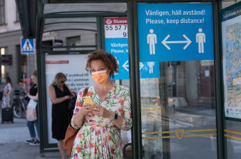 Женщина в маске на автобусной остановке с информационным знаком, призывающим людей сохранять социальную дистанцию из-за пандемии коронавируса COVID-19 в Стокгольме