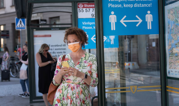 Женщина в маске на автобусной остановке с информационным знаком, призывающим людей сохранять социальную дистанцию из-за пандемии коронавируса COVID-19 в Стокгольме