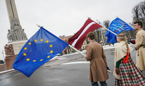 Юноша и девушка в национальных латышских костюмах с флагами ЕС и Латвии у Памятника Свободы в Риге